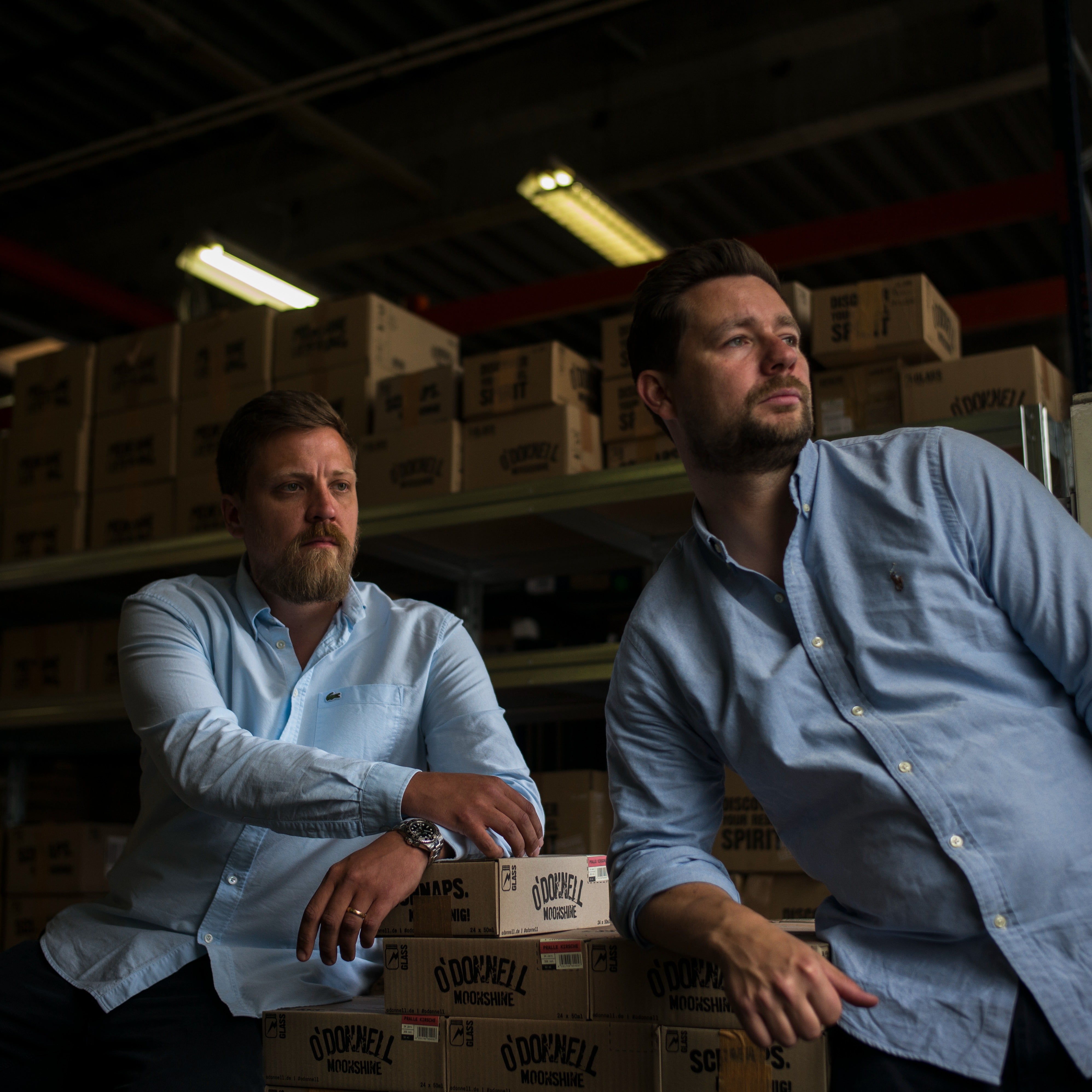 Die beiden Gründer von O'Donnell Moonshine stehen in hellblauen Hemden vor hohen Lagerregalen voller Versandkartons in ihrem Berliner Warenlager.
