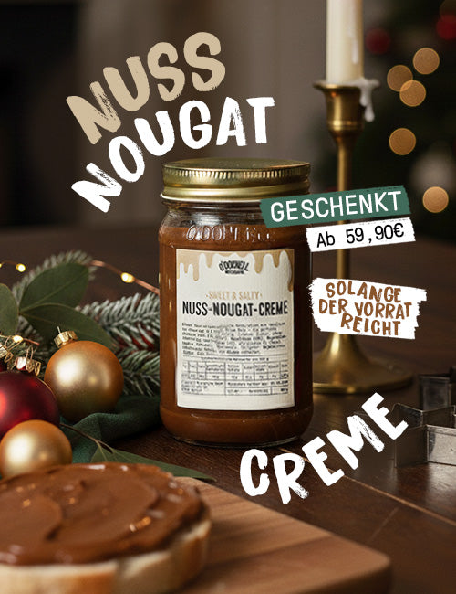 Ein dunkles, warm getöntes Hero-Bild, das ein Glas Nuss-Nougat-Creme auf einem rustikalen Holztisch zeigt, umgeben von Weihnachtsdekorationen wie goldenen Christbaumkugeln und einer brennenden Kerze. Die wichtigsten Text-Overlays lauten: „Nuss Nougat Creme“ und ein Banner, das auf ein Sonderangebot hinweist: „GESCHENKT Ab 59,90€“ und „SOLANGE DER VORRAT REICHT“. Im Vordergrund ist eine Brotscheibe mit dem Aufstrich zu sehen.