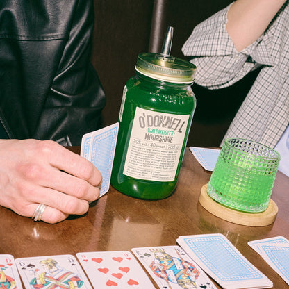 Ein großes, grünes O'Donnell „Waldmeister“ Likörglas mit Ausgießer steht inmitten von Spielkarten und einem Drink auf einem Holztisch.